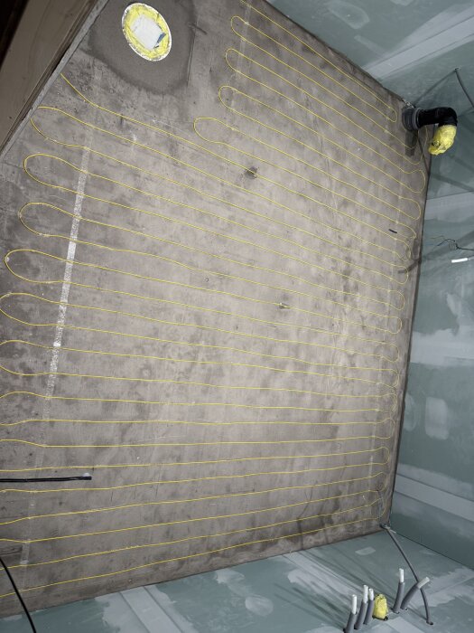 Gul elgolvvärmeslinga monterad på ett betonggolv i ett rum under renovering, med synliga rör och brunnen i vänstra hörnet.