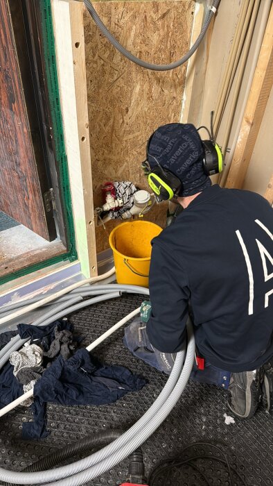 Person installerar rör vid vägg med synlig vattenmätare, iklädd hörselskydd och arbetshandskar, bredvid en gul hink och hoprullade textilier.