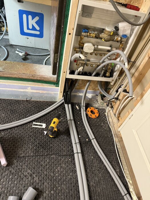 Vattenmätarskåp med rörinstallationer, elverktyg och kablar på ett gummigolv; del i renoveringsprojekt för badrum och garderob.