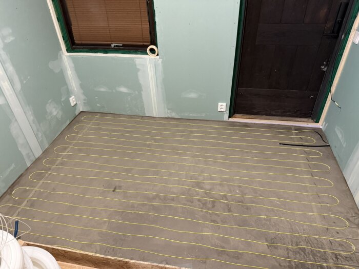 Elgolvvärmeslingor monterade på ett nyligen flytspacklat golv i ett rum under renovering, med gröna väggar och en dörr i mörkt trä.