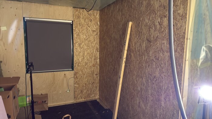 Rum under renovering med OSB-väggar, rullgardin, och verktygslåda. Träregel står lutad mot väggen och lampor ger starkt ljus.