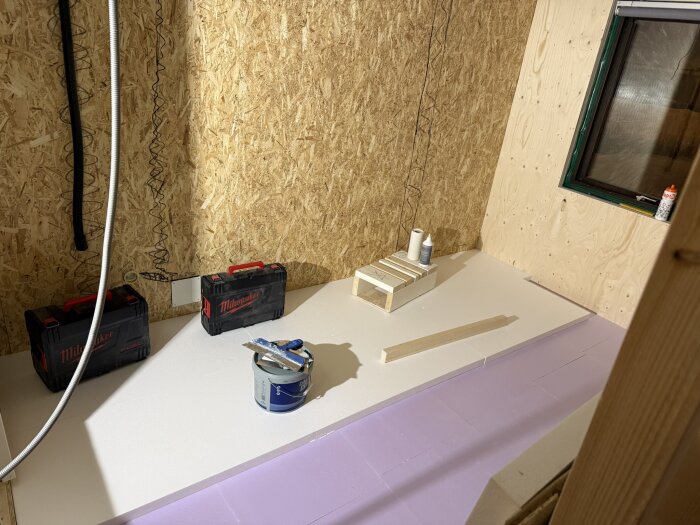 Verktygslådor och byggmaterial på golv i renoveringsprojekt, med trådskivor och ett fönster i bakgrunden.