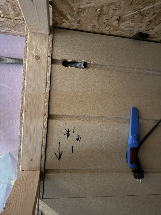 Närbild på en vägg med trä- och spånskivmaterial, markerade pilar och siffror, samt ett blått verktyg bredvid en elkabel.