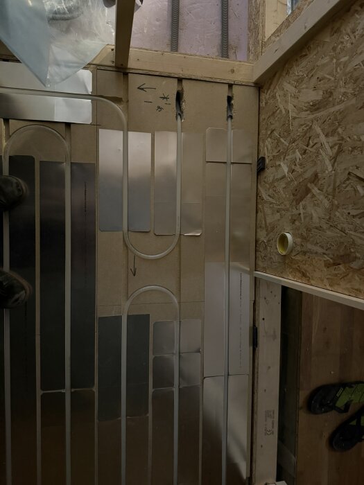 Golvvärmeinstallation med rör och metallplattor, monterat på träpanel mot isolerad vägg. Instruktioner skrivna på ytan för montering.