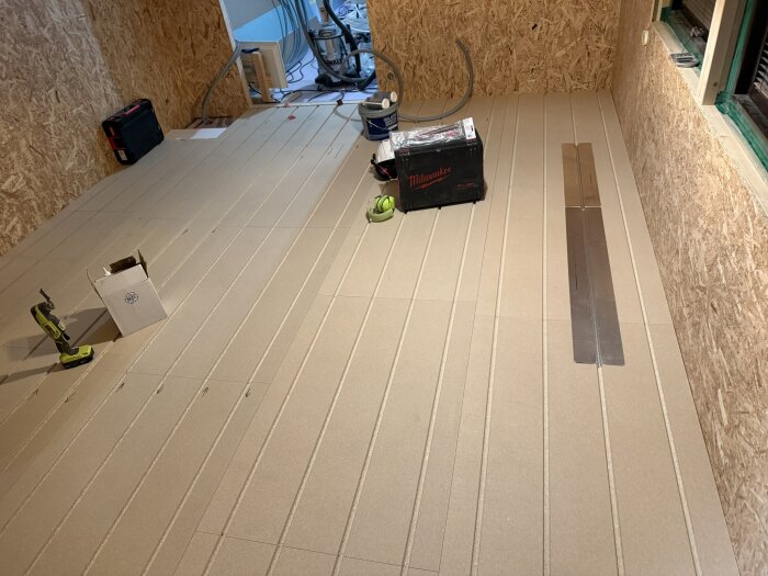 Rum med trägolv under konstruktion, verktyg och utrustning ligger på golvet, inkl. en borrmaskin och en kartong.