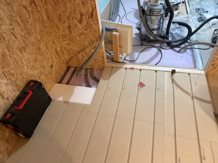 Verktygslåda och dammsugare på ett bygge med isoleringsskivor och rördragningar på golvet i köket under renovering.
