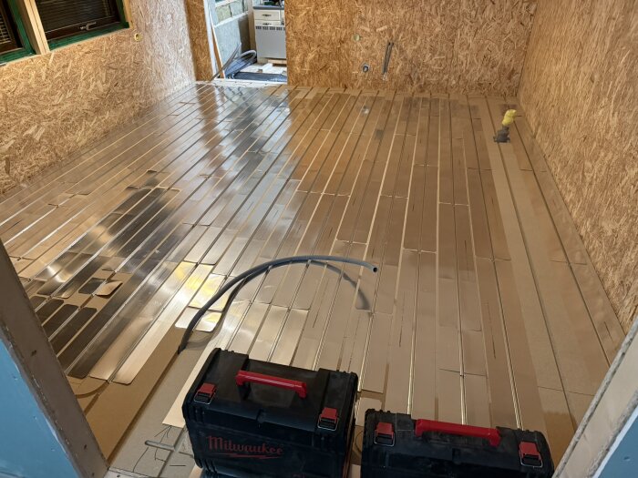 Golvyta med nylagd gulvvärmeinstallation och golvplattor, omgiven av träväggar. Två verktygslådor syns i förgrunden.