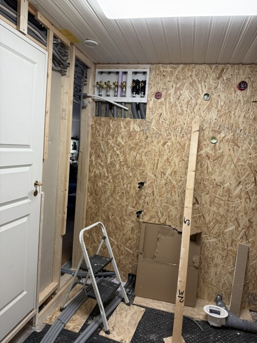 Renovering av rum med synliga rörinstallationer, en stege framför en vägg av spånskiva, flera rör uppsatta längs väggen och även ett ventilationsskåp.