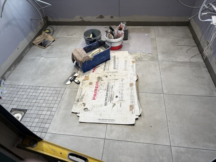 Verktyg och material på ett nylagt klinkergolv i ett rum under renovering, med kartong och byggmaterial i hörnet.