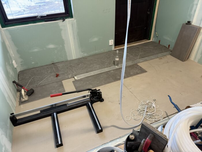 Verktyg och byggmaterial i ett rum under renovering, med oinstallerade plattor och en kakelskärare på golvet.
