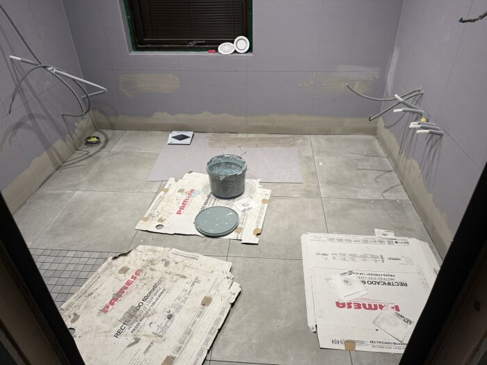 Badrum under renovering med golvplattor, kablar hängande från väggarna och färgburk på golvet, väggarna är delvis spacklade.