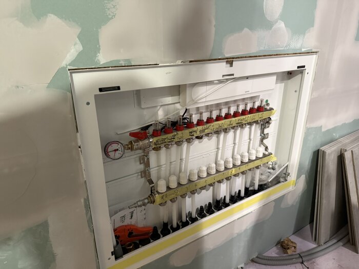 Värmeskåp monterat i vägg, med manifolder och röranslutningar för golvvärmeinstallation, omgiven av grönt gipsskiva och kakelplattor.