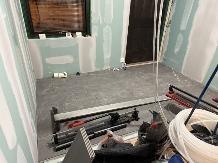 Rum under renovering med nylagt golv, väggar spacklade och verktyg och material utspridda, fönster och dörr synliga.