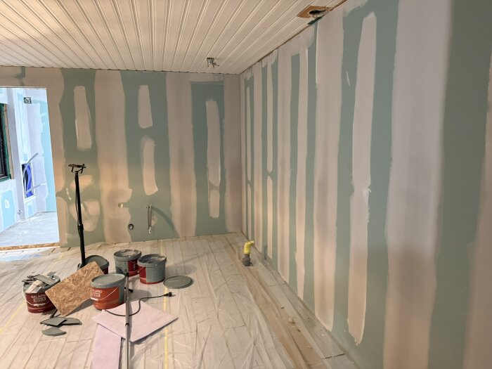 Rum under renovering med spacklade gipsskivor på väggarna, skyddsplast på golvet och målarburkar samt verktyg på golvet.