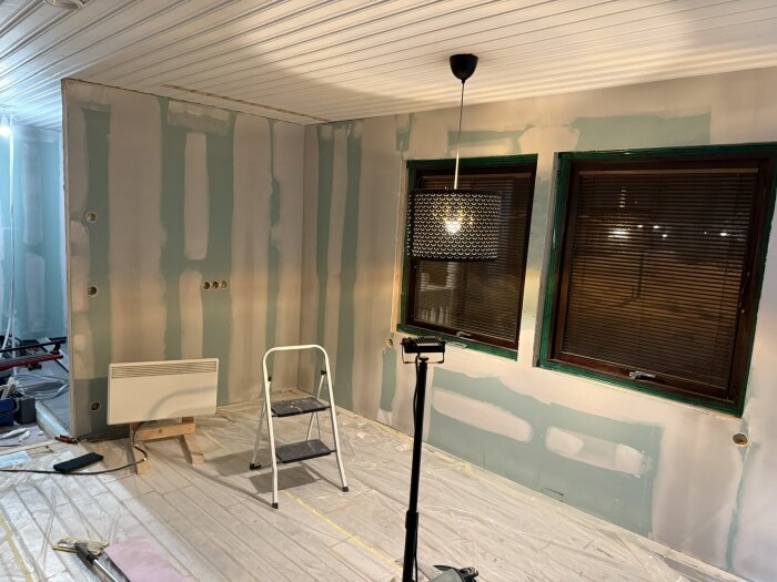 Rum under renovering med spacklade väggar, en stege, värmeelement och lampa. Fönster har bruna rullgardiner och gröna foder.