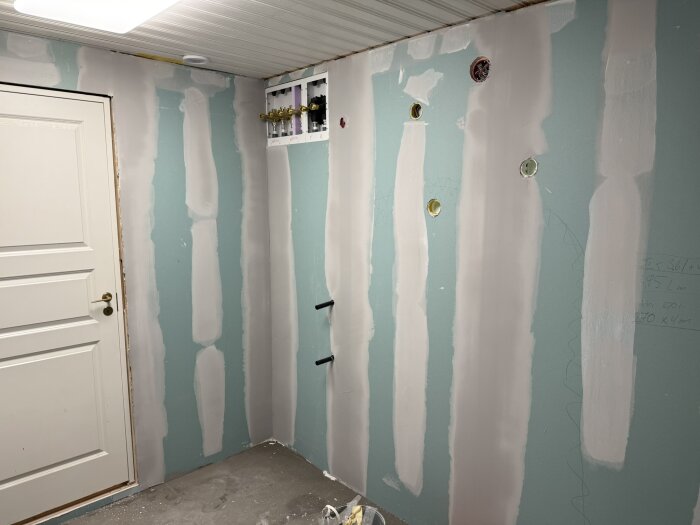 Ett rum under renovering med nyligen spacklade väggar och synliga röruttag, samt en vit dörr till vänster.