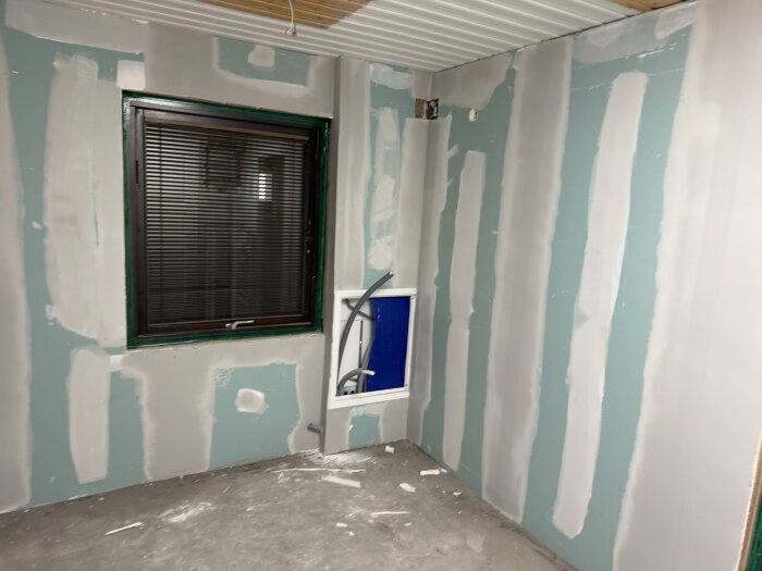 Spacklade väggar med gröna gipsskivor i ett rum under renovering, fönster med persienner och utrymme för vägghängd toalettcistern syns.
