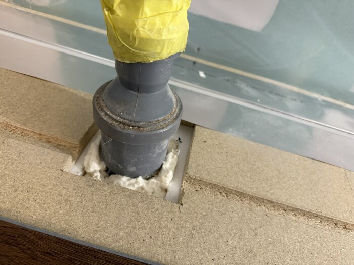 Grått rör i skiva, tätat med fogskum runt basen. Röränden är täckt med gul plast. Bakgrund av trä och vägg i ljus färg.