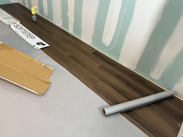 Vinylgolv i köket under installation, med en rulle underlagsmaterial på golvet intill och gipsade väggar i bakgrunden.