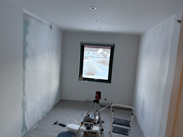 Ett rum under renovering med spacklade väggar, verktyg på golvet, en stege och en laserpass. Ett fönster visar snö utomhus.
