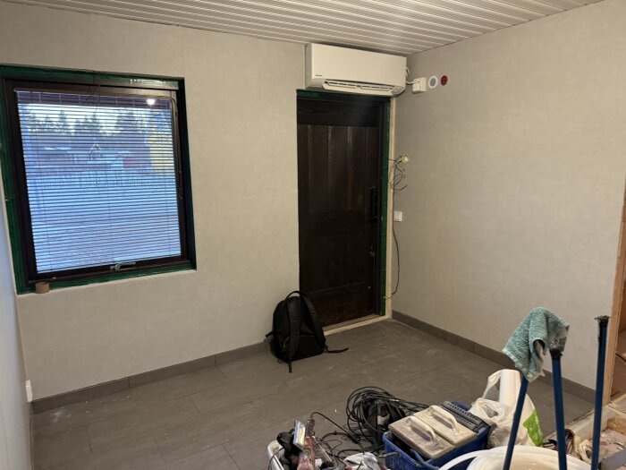 Renoveringsrum med grått golv, väggar och innertak, fönster med persienner, träfärgade dörrar; verktyg och material på golvet.