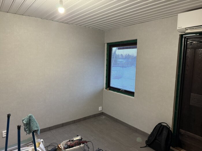 Rum under renovering med nylagt golv och en målad vägg. Ett fönster med persienner, takbelysning och byggverktyg syns på golvet.