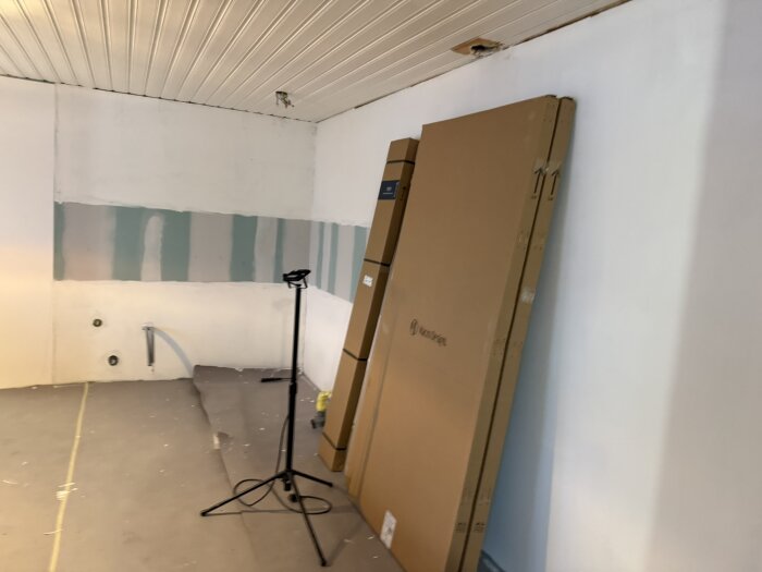 Renovering av kök pågår med obruten vägg, rörutgångar, golvbeläggning, kartonger lutade mot väggen och en mikrofonstativ i förgrunden.