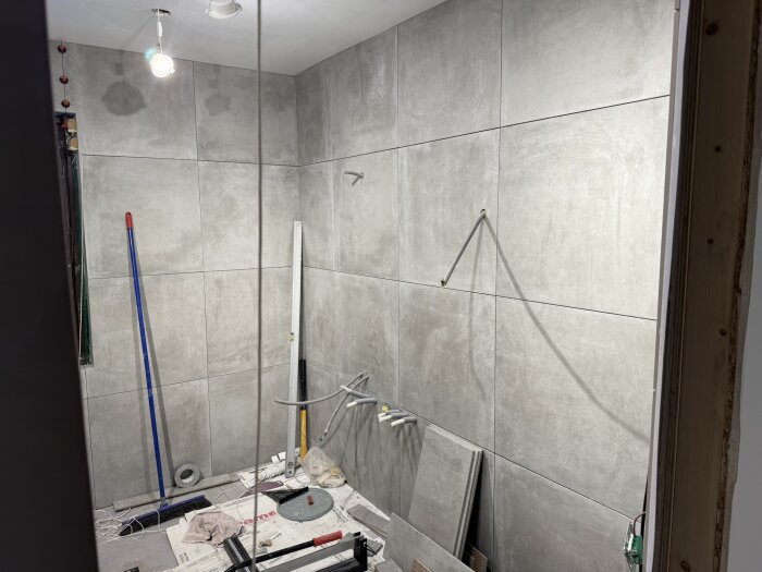 Renovering av badrum med stora grå klinkerplattor på väggarna, verktyg och material ligger på golvet, pågående projekt.