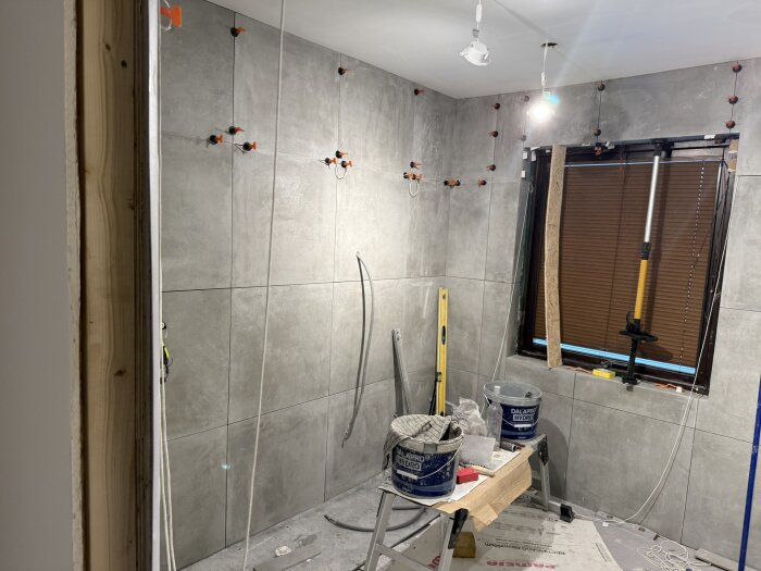 Renovering pågår i ett rum med stora gråa kakelplattor på väggarna, arbetsverktyg och material syns på golvet.