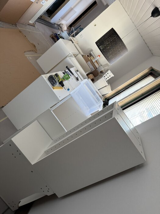 Kök under renovering med halvmonterade köksskåp och verktyg på golvet, ny belysning och öppnade kartonger i bakgrunden.