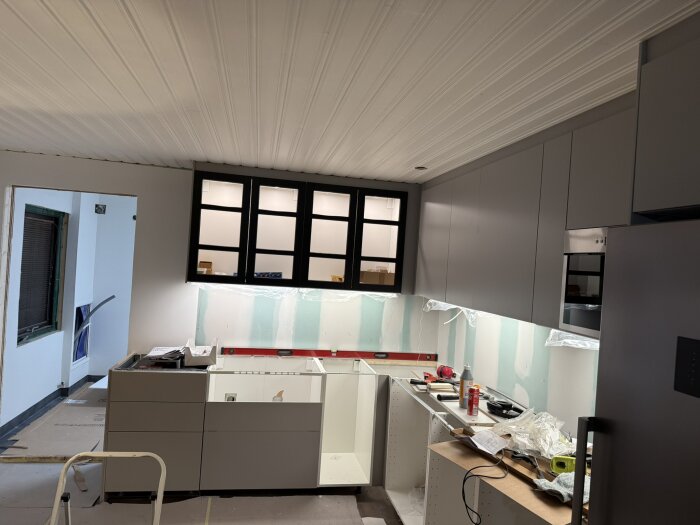 Nytt kök under uppbyggnad med grå skåp från IKEA, en inbyggd ugn, och en del av bänkskivan saknas. Verktyg och material ligger på bänkarna.