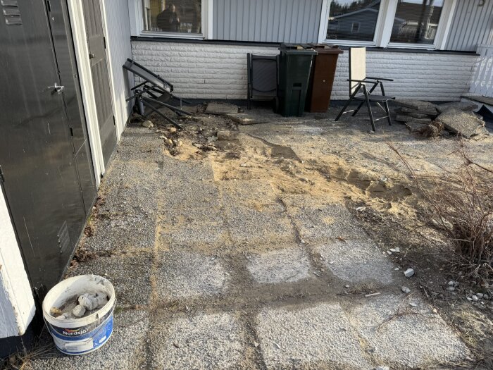 Uteplats med stökig mark, grus, vissna buskar, två sopkärl, hopfälld solstol och hink mot en vitputsad vägg.