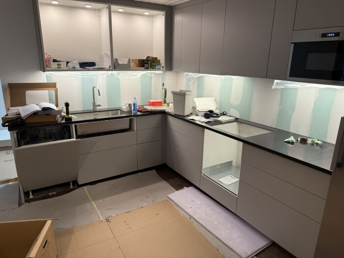 Nytt kök under installation med grå skåp, svart bänkskiva, diskho och öppen diskmaskin. Skåpen har spotlights. Material och verktyg syns på bänken.