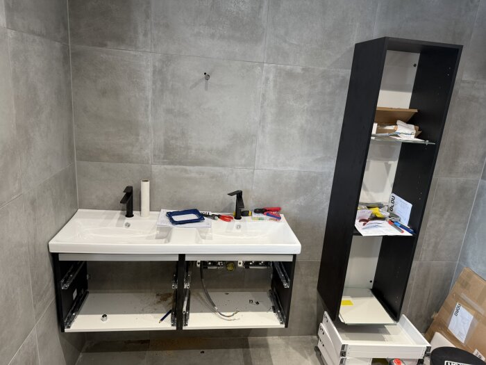Badrum under renovering med dubbelhandfat utan luckor, verktyg och material på bänken samt ett högt svart skåp med öppna hyllor och lådor.