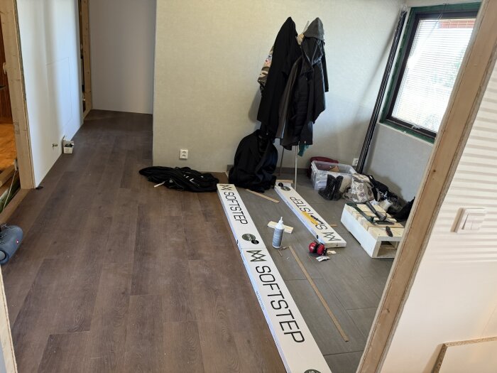 Rum under renovering med mörkt golv, kläder på galge, material kartonger märkta "Softstep," och diverse verktyg och material på golvet.