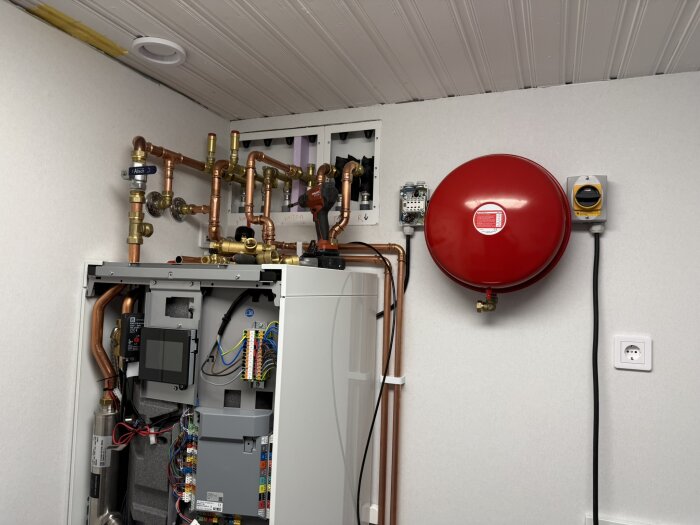 En bergvärmepump med synliga rörkopplingar och en röd tryckbehållare monterad på väggen i ett tekniskt rum.