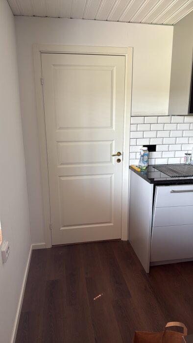 Vit dörr med modernt handtag i rum med mörkt trägolv, synlig köksbänk och del av vit kakelvägg samt rengöringsspray.