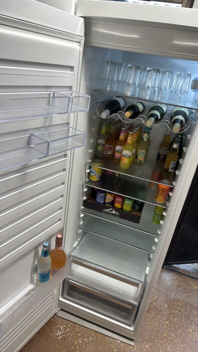 Öppen kyl fylld med flaskor, burkar och glas, inklusive alkoholhaltiga drycker såsom Breezer och Sol, i ett garage.
