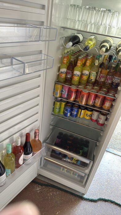 Öppen kyl i garage med flaskor, glas och burkar i flera hyllor. Kylens dörr har vinflaskor placerade i en hylla och golvet är av cement.