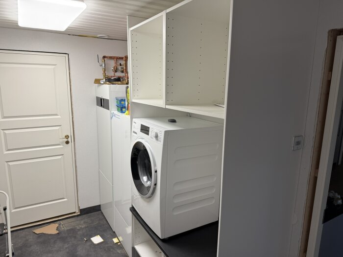 Tvättstuga med vit tvättmaskin och torktumlare på upphöjd plattform, inredning monterad ovanpå, del av dörr och vägg syns också.