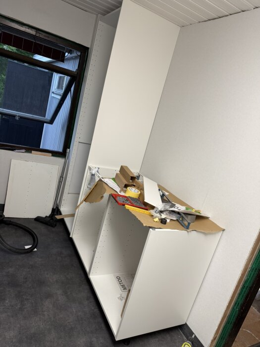 Vit garderobsstomme under montering med verktyg och kartong ovanpå, vid ett fönster i ett renoverat rum.