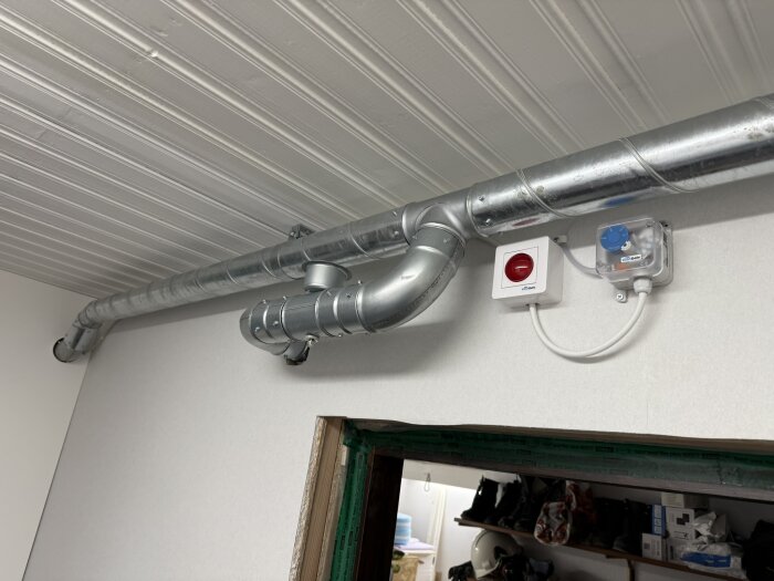 Ventilationsinstallation med metallrör och en kontrollpanel på väggen nära taket i ett garage eller förråd.