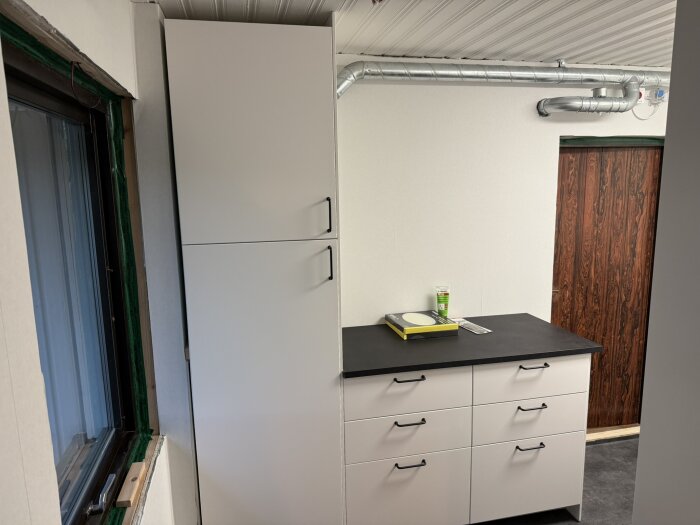 Kök med vit skåpsinredning, svart bänkskiva, fönster och ventilation.