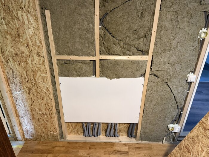 Vägg med isolering och en vit platta fastskruvad, flera grå slangar går in under plattan. Träpanel och OSB-skivor syns på sidan.
