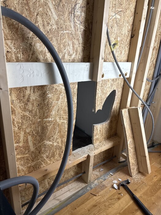 Vägg under renovering med uttaget hål för inbyggd tv-bänk, reglar och dolda slangar för kablar syns. Hammare och spån på golvet.