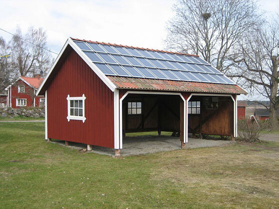 Rödmålat garage med sadeltak och solpaneler, dubbla öppningar, placerat på gräsig tomt, äldre byggnad i bakgrunden.