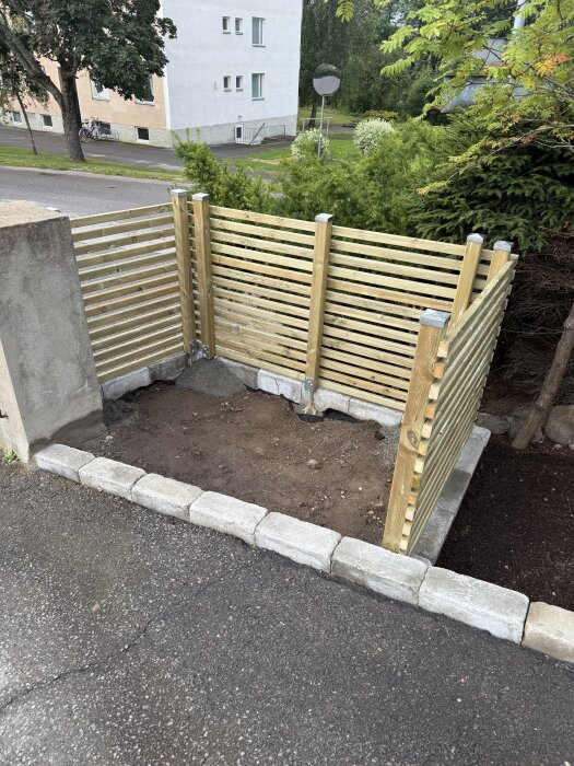 Grusad yta med trästaket runt, där en betongplatta ska gjutas för soptunnor, omgiven av stensättning och buskage i bakgrunden.