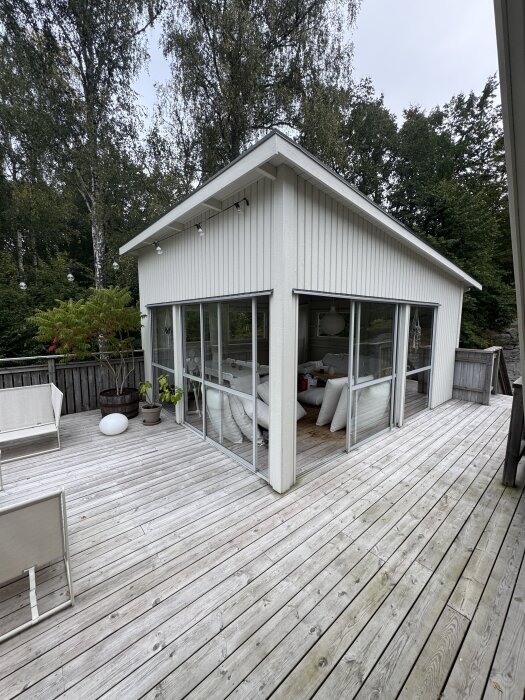 Uterum med stora skjutdörrar öppna, byggt ovanpå ett garage, omgiven av trädäck och träd, med insyn till inredningen och växter utanför.