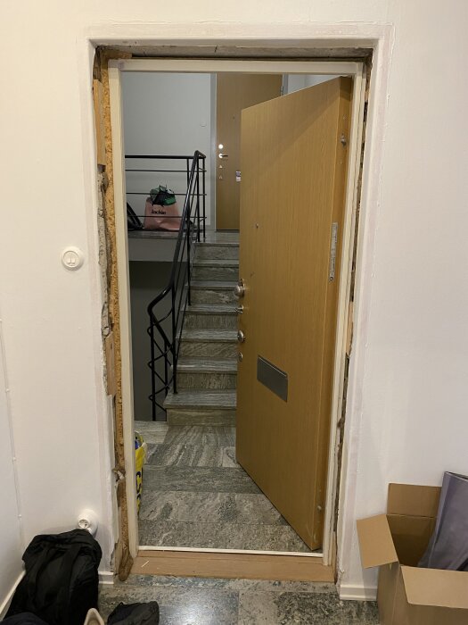 Lägenhetsdörr med synlig puts och ram runt, borttagen plåtklädsel, vy mot trappa och annan dörr i bakgrunden.