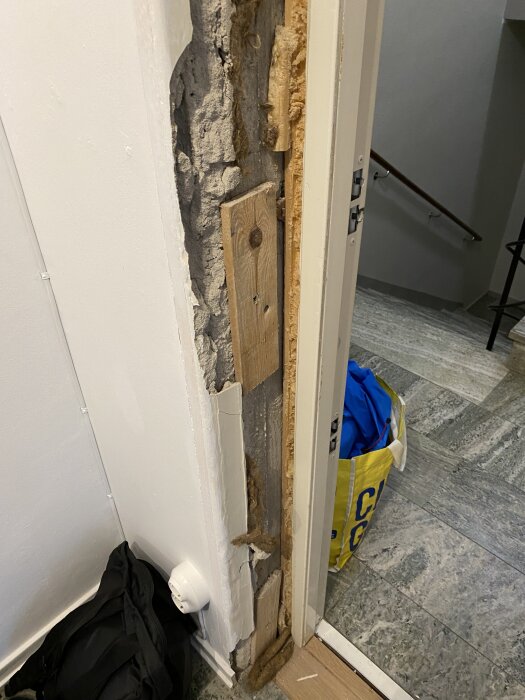 Inre del av dörrkarm utan puts, med synligt trä och isolering vid säkerhetsdörr i lägenhet; Ikea-påse och trappa synliga i bakgrunden.